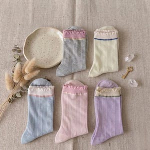 May include: Six pairs of colorful socks with ruffled tops. The socks are in shades of gray, light blue, pink, yellow, and purple. The socks are arranged on a neutral-colored surface with a small plate and decorative elements.