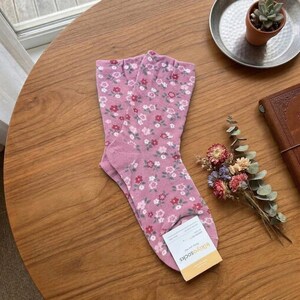 Pode incluir: Um par de meias rosa com um padr&atilde;o floral de pequenas flores vermelhas, brancas e rosa. As meias est&atilde;o dobradas sobre uma mesa de madeira, com uma etiqueta que diz "kikiyosocks". Um pequeno buqu&ecirc; de flores secas e uma suculenta em um vaso tamb&eacute;m est&atilde;o na mesa.