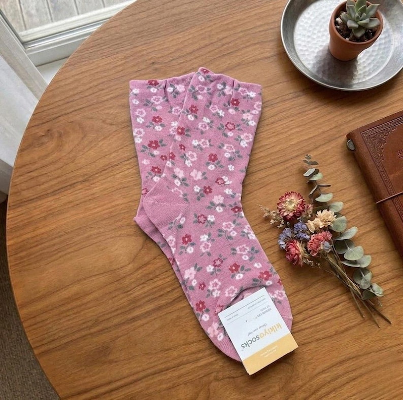 Pode incluir: Um par de meias rosa com um padr&atilde;o floral de pequenas flores vermelhas, brancas e rosa. As meias est&atilde;o dobradas sobre uma mesa de madeira, com uma etiqueta que diz "kikiyosocks". Um pequeno buqu&ecirc; de flores secas e uma suculenta em um vaso tamb&eacute;m est&atilde;o na mesa.