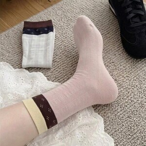 Puede incluir: Un calcetín rosa claro con un puño marrón y beige se lleva en un pie. Un par de calcetines blancos con un puño marrón están doblados cerca. Una zapatilla negra está en el fondo. Los calcetines están sobre una alfombra marrón claro.