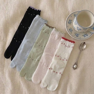 May include: Five pairs of split-toe socks in black, light blue, sage green, pale pink, and white. Each sock features a delicate ruffled trim at the cuff and a subtle dotted pattern. The socks are arranged in a fan shape next to a floral teacup.