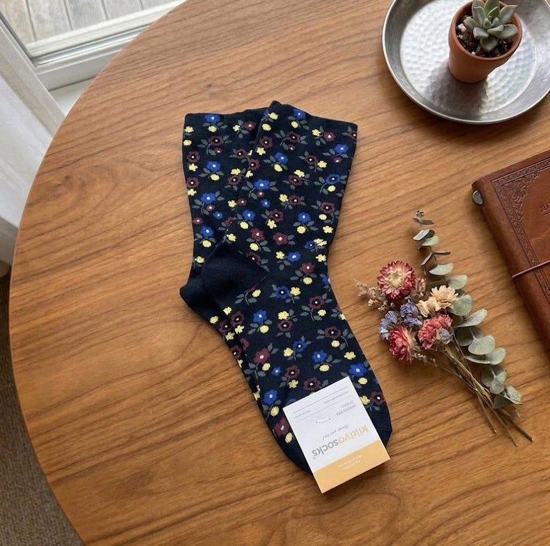 Pode incluir: Um par de meias azul marinho com um padr&atilde;o floral em azul, amarelo e bord&ocirc;. As meias est&atilde;o sobre uma mesa de madeira, com uma pequena etiqueta anexada. Um pequeno buqu&ecirc; de flores secas e uma suculenta em um vaso tamb&eacute;m s&atilde;o vis&iacute;veis.