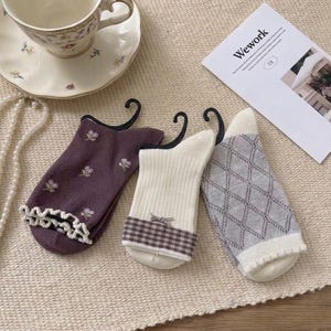 May include: Three pairs of socks with different designs. One is purple with floral embroidery and a ruffled edge. Another is white with a plaid cuff and a bow. The third is gray with a diamond pattern.