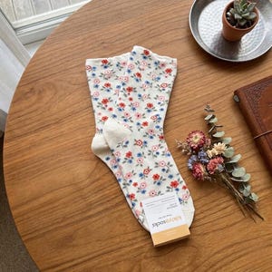 Pode incluir: Um par de meias brancas com um padr&atilde;o floral de flores vermelhas, rosa e azuis. As meias est&atilde;o dobradas sobre uma mesa de madeira, com uma pequena etiqueta anexada. As meias s&atilde;o da marca kikiyasocks.