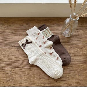Könnte beinhalten: Drei Paar Socken in Creme, Braun und Weiß. Ein Paar hat eine braune Schleife, ein anderes ein Blumenmuster und das dritte einen grün-weißen Besatz. Die Socken liegen auf einer Holzoberfläche.