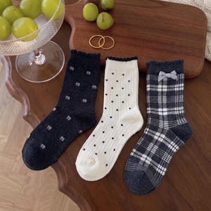 May include: Three pairs of crew socks with ruffled cuffs. The first is dark gray with small white floral patterns. The second is cream with black vertical stripes and dots. The third is a dark gray and white plaid pattern with a small gray bow.
