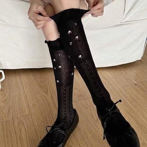 May include: Black knee-high socks featuring a delicate white floral pattern and vertical ribbed detailing along the lower leg, paired with black leather ballet flats that have a rounded toe and thin laces tied across the top of the foot.