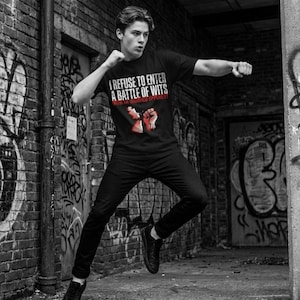 May include: Black t-shirt with the text "I REFUSE TO ENTER A BATTLE OF WITS WITH AN UNARMED OPPONENT" in white and red. The shirt features a graphic of a red fist. The person is wearing black trousers and shoes.