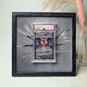 May include: A black framed display featuring a PSA graded 2023 Pokemon Charizard ex Special Illustration Rare card. The card is mounted on a gray background with a white and black paint splatter effect. A clear glass vase holds a pink pampas grass stem.