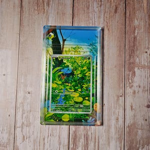 Op de afbeelding: Een Bulbasaur-verzamelkaart in een doorzichtige plastic beschermhoes met een bedrukte bosachtergrond. De kaart toont de naam Bulbasaur, 70 HP en de tekst voor de Leech Seed-aanval. De afbeelding toont een blauw wezen in groen gebladerte.