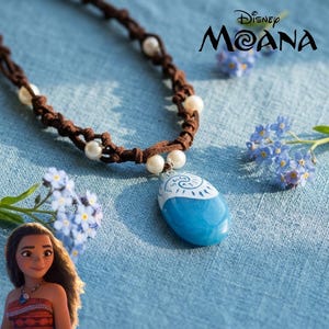 Pode incluir: Um colar de cordão castanho com contas brancas e um pendente oval azul com um design de espiral branco. O pendente é inspirado na personagem da Disney Moana, que é retratada no canto com um top estampado e um colar semelhante.