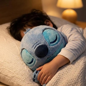 Puede incluir: Un peluche de Stitch azul claro con ojos grandes y cerrados y una nariz azul oscuro. El peluche está siendo abrazado por una persona que lleva una camisa de manga larga a rayas, descansando sobre una almohada blanca texturizada.