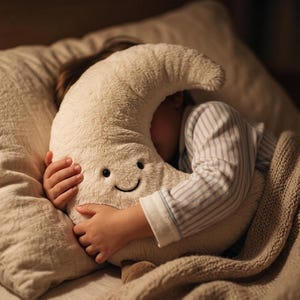 Puede incluir: Una almohada de felpa en forma de media luna, de color crema, con una cara sonriente, es sostenida por una persona que lleva una camiseta de pijama a rayas. La almohada está metida contra una almohada y una manta, creando una escena acogedora.