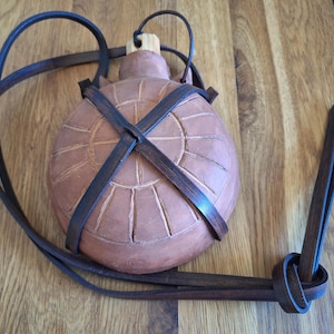 Peut inclure: Une gourde en céramique marron avec une sangle et des accents en cuir. La gourde a une forme ronde avec un motif de soleil sculpté. La sangle en cuir est marron foncé et enveloppe la gourde. La gourde mesure environ 18 cm de hauteur.