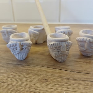 May include: A group of small, unglazed ceramic pipes with carved faces and a wooden stem. The pipes are arranged on a wooden surface, with a white tiled background. The faces have detailed features, including eyes, a nose, and a beard.