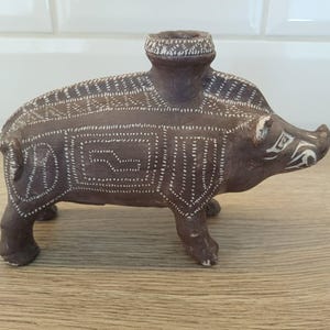 Puede incluir: Figura de cerámica marrón de un jabalí con patrones geométricos blancos. El jabalí tiene una abertura cilíndrica en la espalda y está sobre cuatro patas. Se encuentra sobre una superficie de madera.