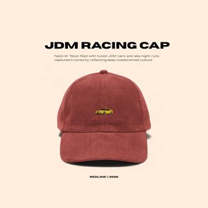 May include: A maroon corduroy JDM Racing Cap with a small yellow car embroidered on the front. The cap features the text "JDM RACING CAP" and "REDLINE © 2026". The design reflects street culture.