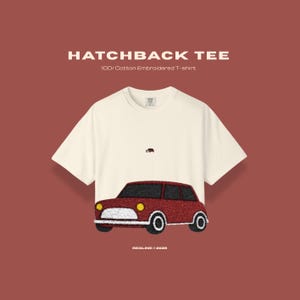 May include: Off-white t-shirt with an embroidered red car design. The shirt has the text "HATCHBACK TEE" and "100% Cotton Embroidered T-shirt". A small car is embroidered above the larger car design.