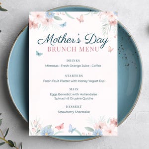Peut inclure: Un menu de brunch pour la fête des mères avec des motifs floraux et papillons. Le menu est sur fond blanc et comprend le texte "Mother's Day Brunch Menu". La carte est posée sur une assiette bleue.