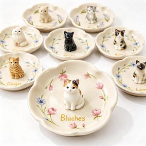 May include: A collection of eight small, round, scalloped-edge ceramic dishes, each featuring a hand-painted floral pattern and a unique three-dimensional cat figurine in the center. Each dish is labeled with a name such as Bluches, Ginger, and Shadow.