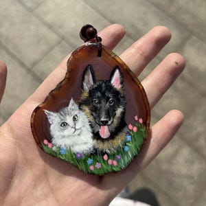 Puede incluir: Retrato de mascota pintado a mano sobre una rodaja de ágata marrón. La obra presenta un perro pastor alemán y un gato blanco y gris, sobre un fondo de hierba verde y flores coloridas. Una cuerda marrón oscuro está unida en la parte superior.