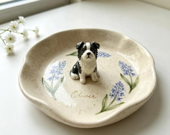 May include: A round, cream-colored ceramic trinket dish with a speckled finish, featuring hand-painted blue floral sprigs and the name Oliver written in cursive. A small, three-dimensional black and white dog figurine sits in the center.