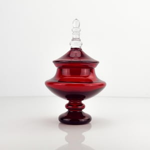 Op de afbeelding: Een dieprode glazen snoeppot met een brede, ronde kom en een bijpassend deksel. Het deksel is voorzien van een heldere, getrapte knop als handvat aan de bovenzijde. Het geheel rust op een ronde voet en heeft een gladde, gepolijste afwerking.