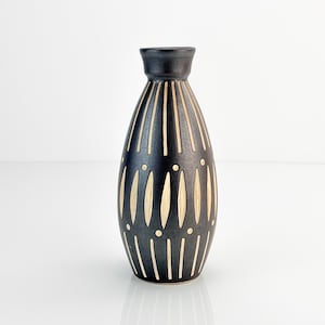 Può includere: Un vaso in ceramica nero opaco con collo stretto e corpo arrotondato. La superficie presenta un motivo ripetitivo di linee verticali, piccoli punti e forme ovali allungate in un colore crema chiaro, creando un design geometrico.