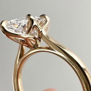May include: A gold engagement ring with a large, clear, pear-shaped diamond. The ring has a delicate, modern design with a thin band and a four-prong setting. The ring is held up by a person's hand.
