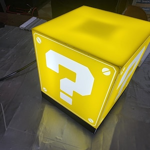 May include: A yellow, illuminated cube with a large white question mark on the front and sides. The cube has a black base and is lit from within. The corners are detailed with white screws. This item is reminiscent of a video game.