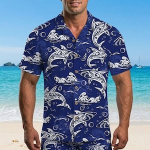 May include: A short-sleeved, button-down shirt in a deep blue color, featuring a repeating pattern of white tribal shark designs, waves, and bubbles. The shirt has a classic collar and wooden buttons. The background shows a beach and ocean.