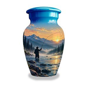May include: A cylindrical cremation urn with a bright blue lid and a glossy finish. The body of the urn features a printed landscape scene of a person fly fishing in a river at sunrise, with tall pine trees, rocky shores, and distant mountains.