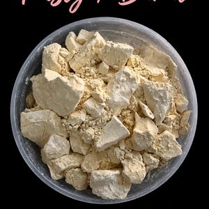May include: A clear plastic container filled with irregular, pale yellow, chalky clay chunks and powder. The text 'Toasty / Burnt' is printed in a pink script font above the container against a solid black background.