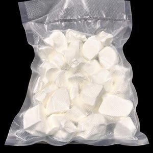 Puede incluir: Una bolsa de plástico transparente llena de numerosos objetos blancos de forma irregular. La bolsa está sellada y parece estar envasada al vacío. El fondo es negro liso, resaltando el contenido.