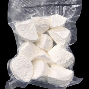 Puede incluir: Un paquete de plástico transparente lleno de trozos de queso blanco de forma irregular. El queso parece blando y tiene una superficie texturizada. El fondo es negro.