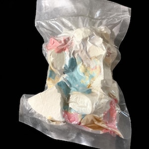 Puede incluir: Una bolsa de plástico transparente y sellada que contiene una variedad de objetos coloridos de forma irregular. Los objetos parecen ser blancos, rosas, azules y amarillos, con una apariencia texturizada. La bolsa está sobre un fondo negro.