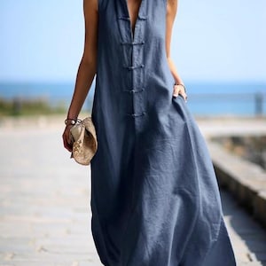 May include: A sleeveless, navy blue maxi dress with a mandarin collar and a series of fabric-covered buttons down the front. The dress has pockets and a flowing skirt. The background shows a beach and blue sky.