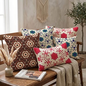 May include: A collection of four square and rectangular throw pillows featuring intricate geometric patterns in shades of dark brown, cream, deep red, and blue, arranged on a wooden bench alongside a book and a small vase with dried grass.