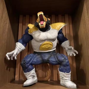 May include: A detailed action figure of a large ape-like character with an open mouth, showcasing sharp teeth. The figure has a white and gold chest plate, blue trousers, and white boots. The figure is in a crouching position.