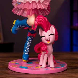 Op de afbeelding: Een verzamelfiguur van een roze pony met krullende manen, staand naast een been met een blauwe veterlaars en een roze rok met ruches. Het figuur staat op een ronde roze voet die is versierd met kleurrijke patronen van ballonnen en confetti.