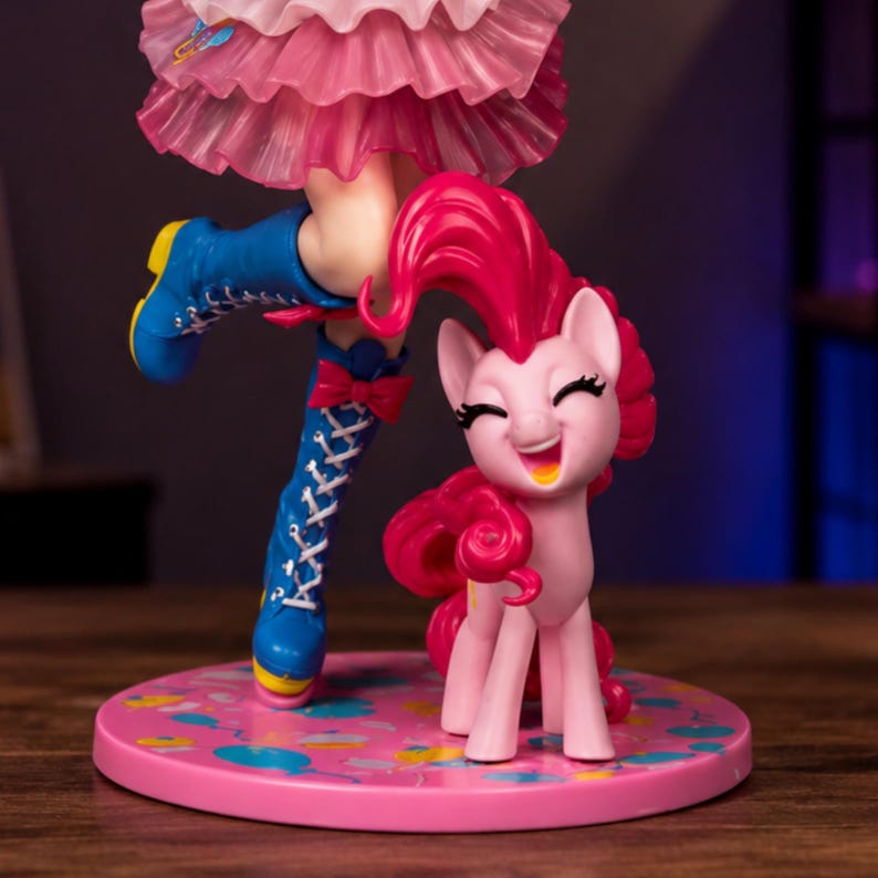 Op de afbeelding: Een verzamelfiguur van een roze pony met krullende manen, staand naast een been met een blauwe veterlaars en een roze rok met ruches. Het figuur staat op een ronde roze voet die is versierd met kleurrijke patronen van ballonnen en confetti.