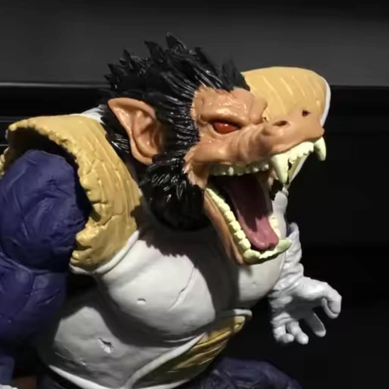 May include: A detailed figurine of a Saiyan character in a monstrous ape form. The figure features a tan face with sharp teeth, red eyes, and black fur. The character wears white and blue armour with gold accents.