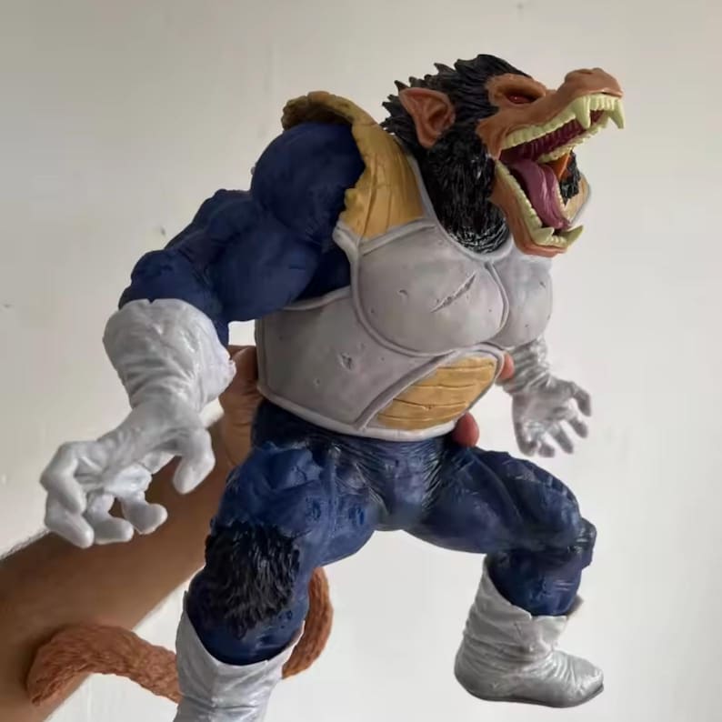 May include: A large figurine of a character in a dynamic pose. The figure has a monkey-like face with an open mouth, revealing sharp teeth and a pink tongue. It wears blue and white armour with white gloves and boots. The figure is held by a person.