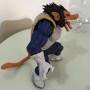 May include: A detailed action figure of a Saiyan character in a battle stance. The figure has a monkey-like face with an open mouth, sharp teeth, and a dark fur mane. It wears blue armour with white gloves and boots, and has a long, orange tail.