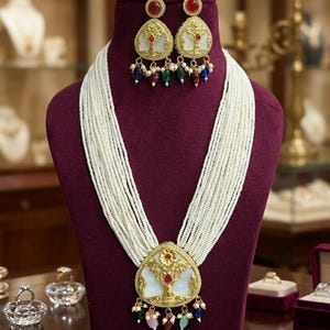 May include: An ornate necklace and earring set displayed on a maroon bust. The necklace features multiple strands of white beads, a gold pendant with a white inlay, and colorful gemstones. The earrings mirror the pendant design.