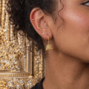 Könnte beinhalten: Nahaufnahme eines Ohrs mit einem goldenen Ohrring. Der Ohrring hat ein glockenförmiges Design mit detaillierten Mustern. Der Hintergrund ist ein verschwommenes, verziertes Goldmuster.