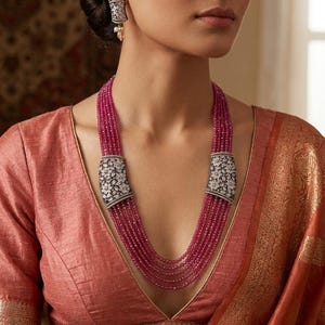Puede incluir: Un collar de rubíes de múltiples hilos con cierres plateados ornamentados, adornado con un patrón floral. También se ven pendientes a juego con un diseño similar. La mujer lleva una blusa color melocotón y un sari naranja estampado.