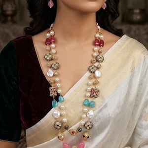 Peut inclure: Un collier multi-rangs composé de perles blanches, de perles translucides roses et bleues, et d'entretoises en métal doré ornées de pierres colorées. Des boucles d'oreilles assorties avec des perles roses et des détails dorés complètent l'ensemble.