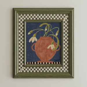 Può includere: Un dipinto incorniciato di fiori bianchi in un vaso di terracotta su sfondo blu scuro. Il dipinto è in una cornice verde con un bordo a scacchi bianco e nero. L'opera d'arte ha uno stile rustico e popolare.