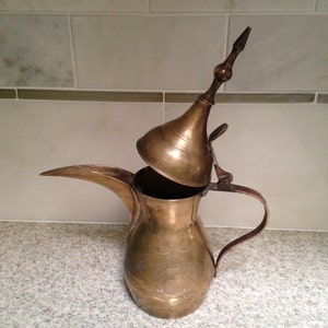 May include: A vintage brass coffee pot with a long spout and a hinged lid. The pot has an ornate design etched into the body and a curved handle. The pot is sitting on a speckled countertop.